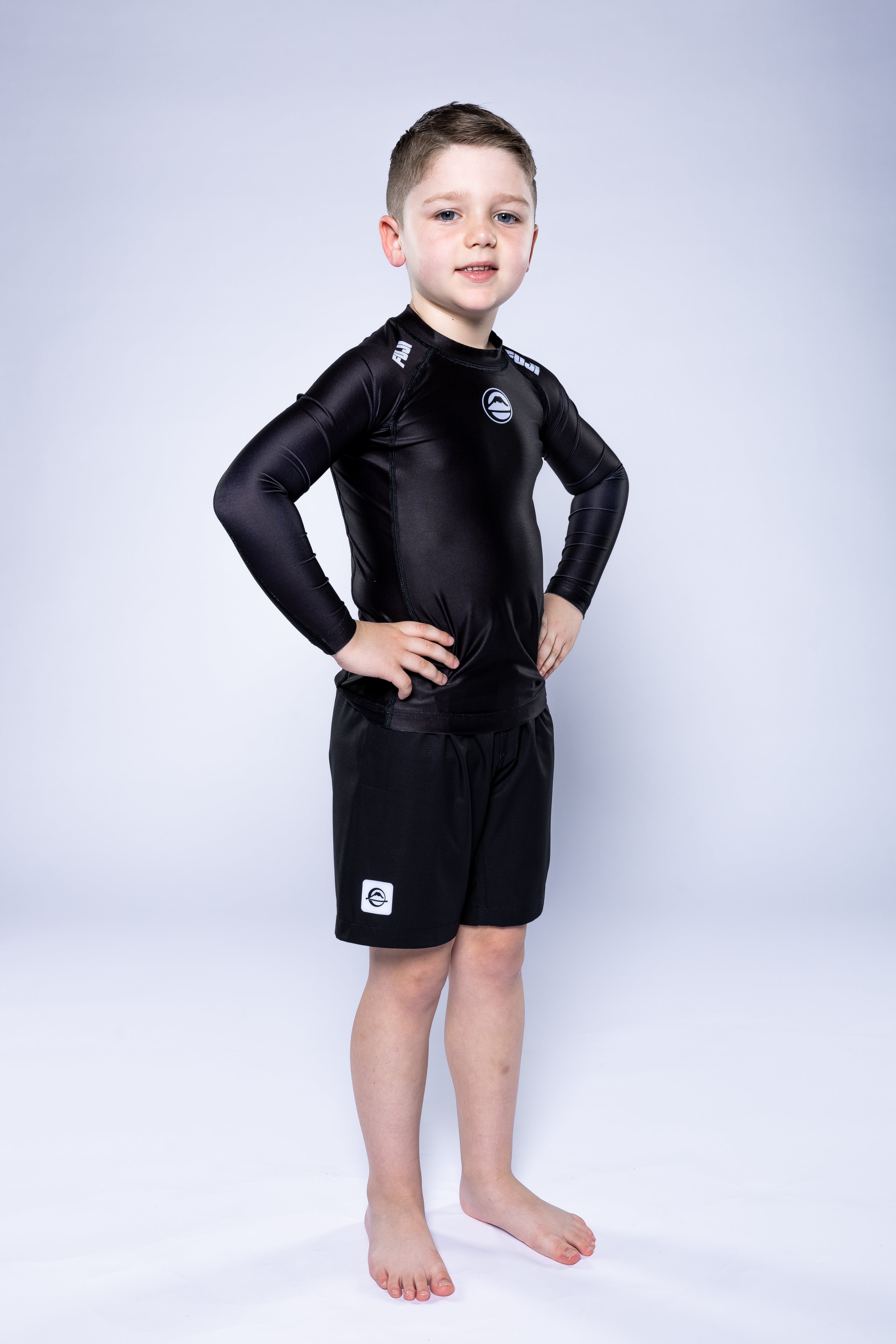A young boy stands barefoot with hands on hips, confidently posing in the Kids Baseline 2.0 Long Sleeve Rashguard Black against a plain light gray background.