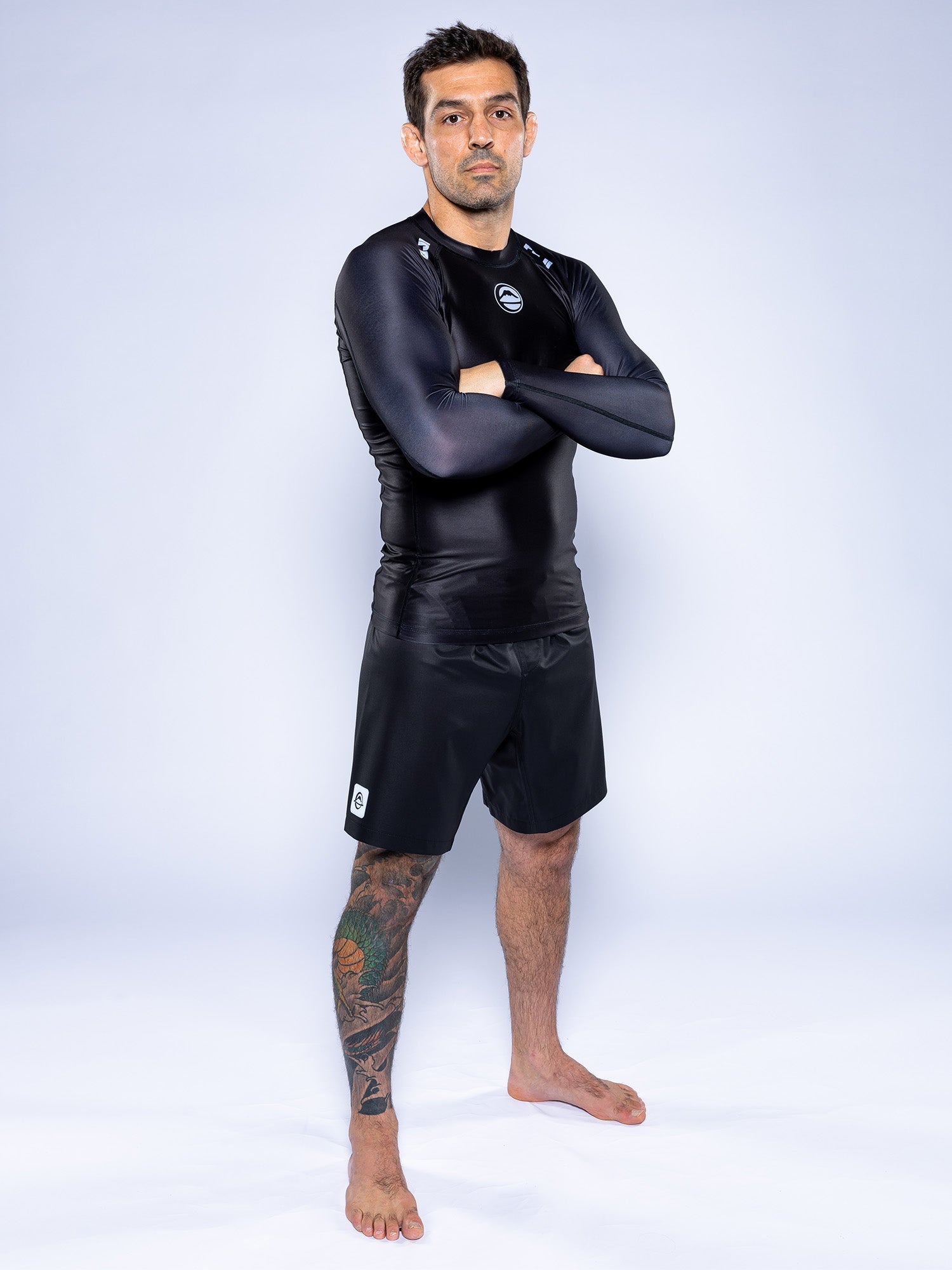 A man with a tattoo on his left leg and a neutral expression stands barefoot, arms crossed, in the Baseline 2.0 Long Sleeve Rashguard Black and black shorts against a plain light background.