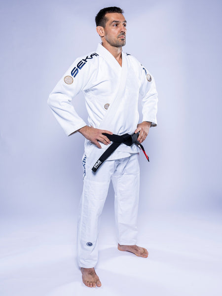 A man in a SEKAI 3.0 BJJ Gi White Navy and a black belt stands barefoot with hands on hips, gazing to the side against a light background.