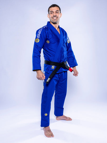 A person stands barefoot on a white background, smiling with arms relaxed at their sides, wearing the SEKAI 3.0 BJJ Gi Blue Yellow and a black belt.