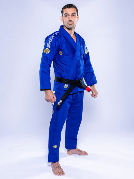 A man stands barefoot on a white background, confidently wearing the SEKAI 3.0 BJJ Gi Blue Yellow with a black belt featuring a red bar, looking at the camera.