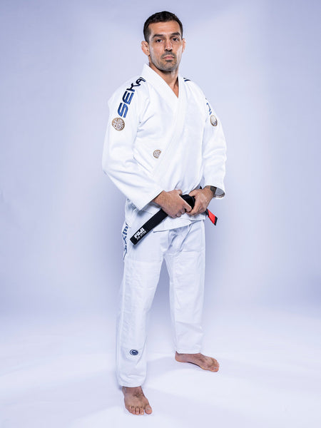 A man in the SEKAI 3.0 BJJ Gi White Navy stands barefoot on a white background, confidently holding a black belt with both hands and looking at the camera.