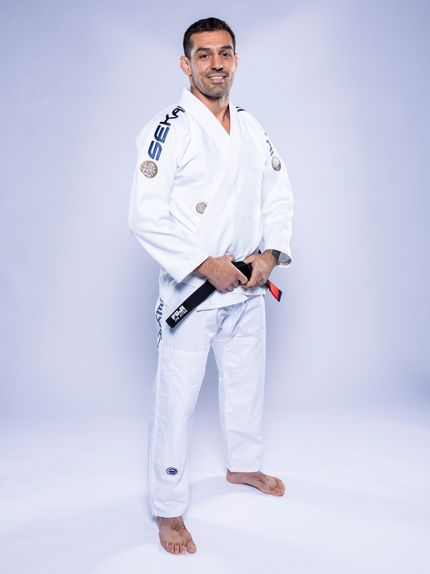 A man wearing the SEKAI 3.0 BJJ Gi White Navy and a black belt stands barefoot on a light background, smiling and holding his belt with both hands.