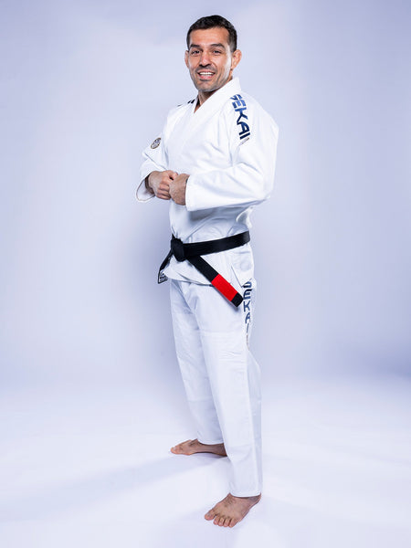A man stands barefoot on a light background, smiling with his hands clasped at his waist, wearing the SEKAI 3.0 BJJ Gi White Navy and a black belt.