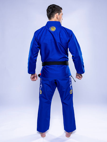 A person wearing the SEKAI 3.0 BJJ Gi Blue Yellow with a black belt stands barefoot, facing away to display the back of the uniform, which features patches and logos on both the jacket and pants.