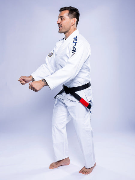 A man stands barefoot on a light background, side-on with arms bent as if preparing for movement, wearing the SEKAI 3.0 BJJ Gi White Navy and a black belt.