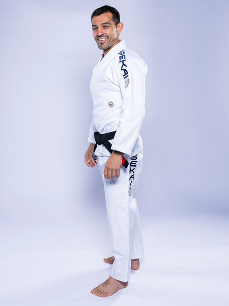 A man wearing the SEKAI 3.0 BJJ Gi White Navy with dark lettering on the sleeve and pants stands barefoot on a white background, smiling and looking at the camera.