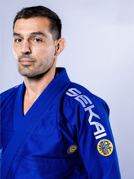 A man models the SEKAI 3.0 BJJ Gi Blue Yellow, featuring SEKAI branding on the shoulder, as he stands against a light background and looks seriously at the camera.