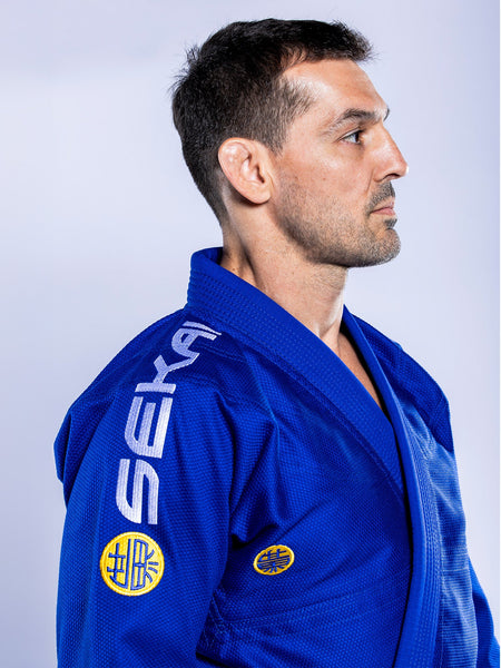 A man wearing the SEKAI 3.0 BJJ Gi Blue Yellow with “SEKAI” on the shoulder and a yellow sleeve patch stands in profile against a light background, facing right.