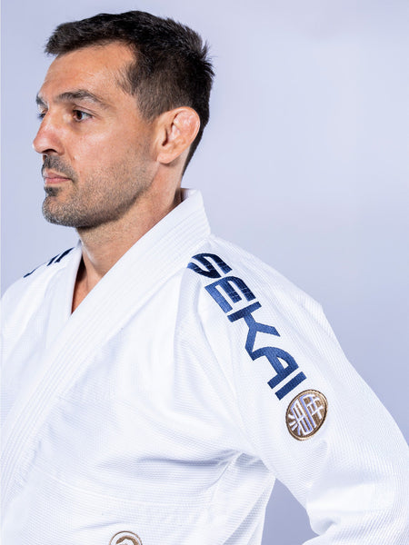 A man with short dark hair and a trimmed beard wears the white-navy SEKAI 3.0 BJJ Gi, embroidered with "SEKAI" on the shoulder, looking left against a plain light background.