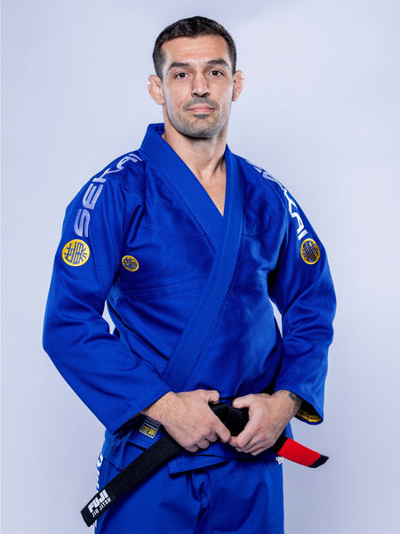A man wearing the SEKAI 3.0 BJJ Gi Blue Yellow with patches stands facing forward, holding a black belt with a red stripe, against a plain light background.