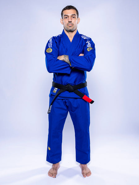 A man wearing the SEKAI 3.0 BJJ Gi Blue Yellow with a black belt stands barefoot on a white background, arms crossed and looking straight ahead.