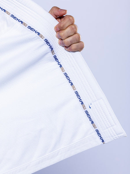 A hand holds the collar of a SEKAI 3.0 BJJ Gi White Navy martial arts uniform, which features the brand name repeatedly printed along the inner edge, against a light purple background.