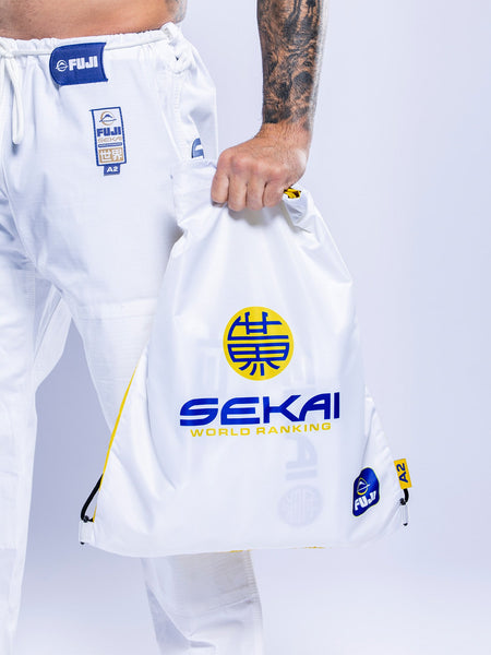 A person in white SEKAI 3.0 BJJ Gi (White Navy) holds a white drawstring bag labeled SEKAI WORLD RANKING with blue, yellow accents, and Japanese characters against a light background.