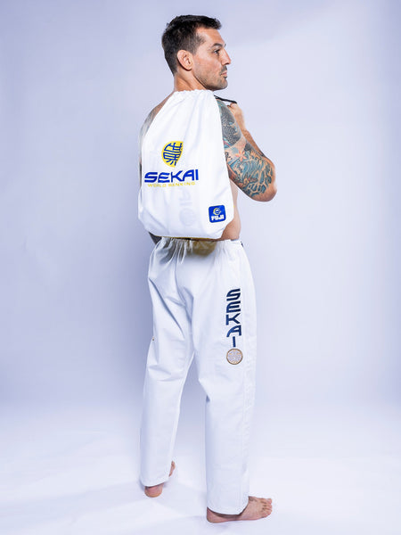 A man in the SEKAI 3.0 BJJ Gi White Navy stands sideways, carrying a matching SEKAI-branded white drawstring bag on his back. Tattoos cover his left arm and he wears a focused expression.