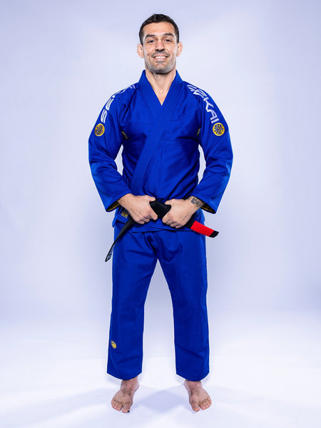 A man stands barefoot on a white background, wearing the SEKAI 3.0 BJJ Gi Blue Yellow and holding a black belt with a red bar while smiling at the camera.
