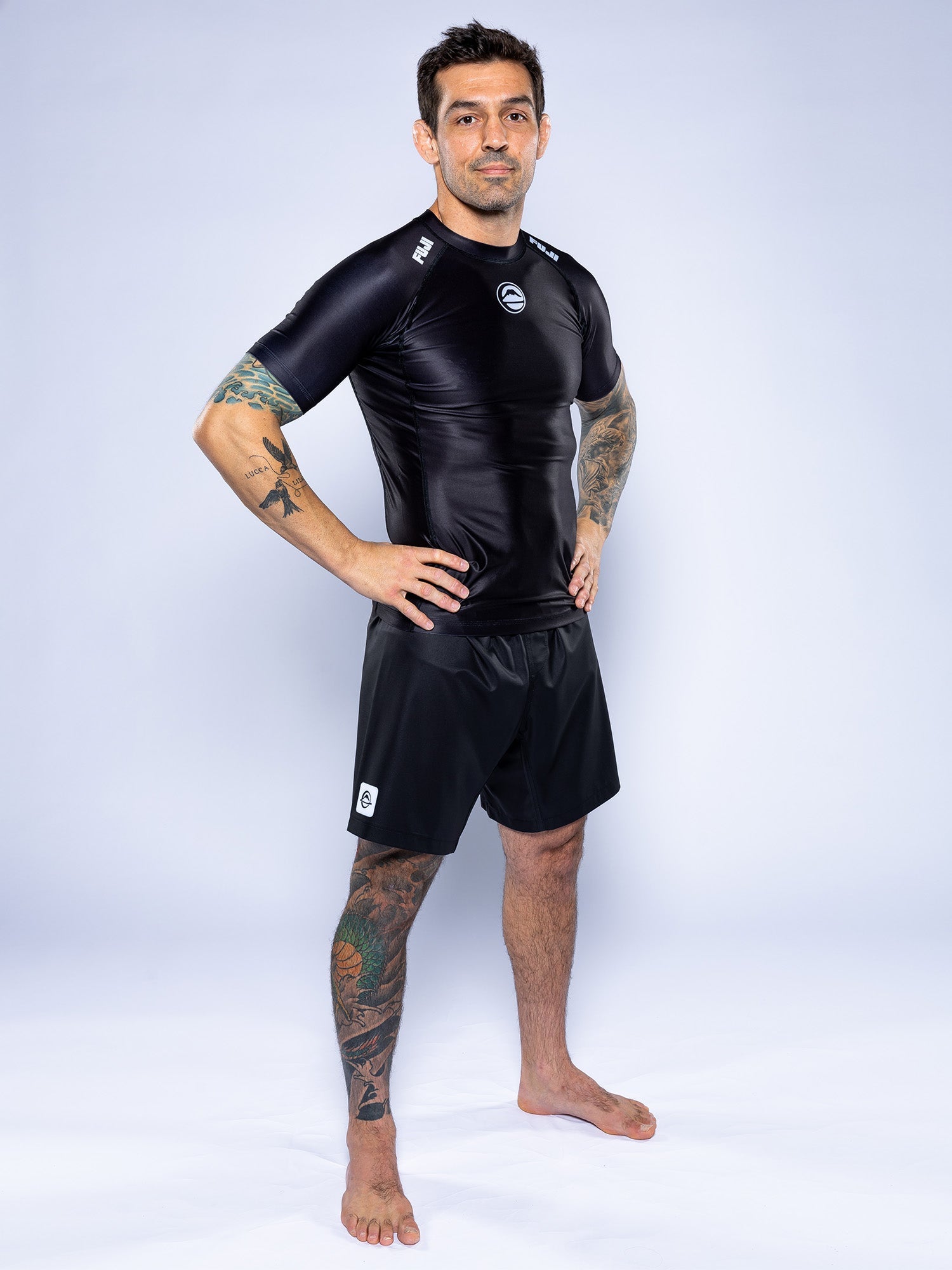 A man with short dark hair and tattoos stands barefoot on a white background, confidently posing in the Baseline 2.0 Short Sleeve Rashguard Black and black shorts, hands on his hips.