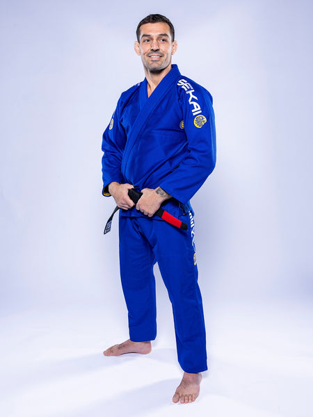 A man wearing the SEKAI 3.0 BJJ Gi Blue Yellow with a black belt stands barefoot on a white background, smiling and facing the camera.