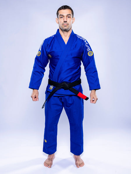 A man stands barefoot on a white background, wearing the SEKAI 3.0 BJJ Gi Blue Yellow with a black belt, facing forward and arms relaxed at his sides.